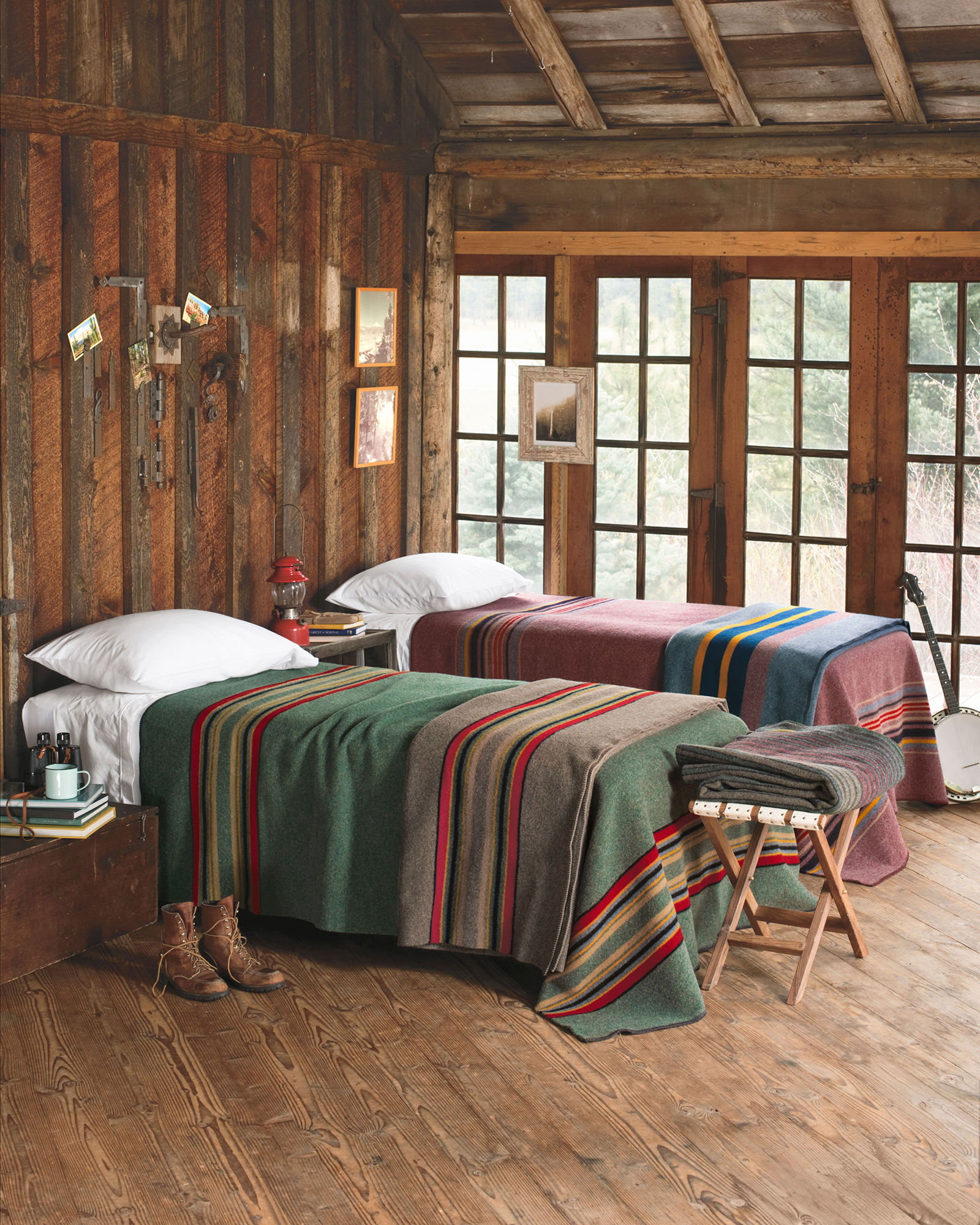 two beds in a log building by a window. Beds are covered with Pendleton Yakima Camp blankets, one green with stripes, one red with stripes.