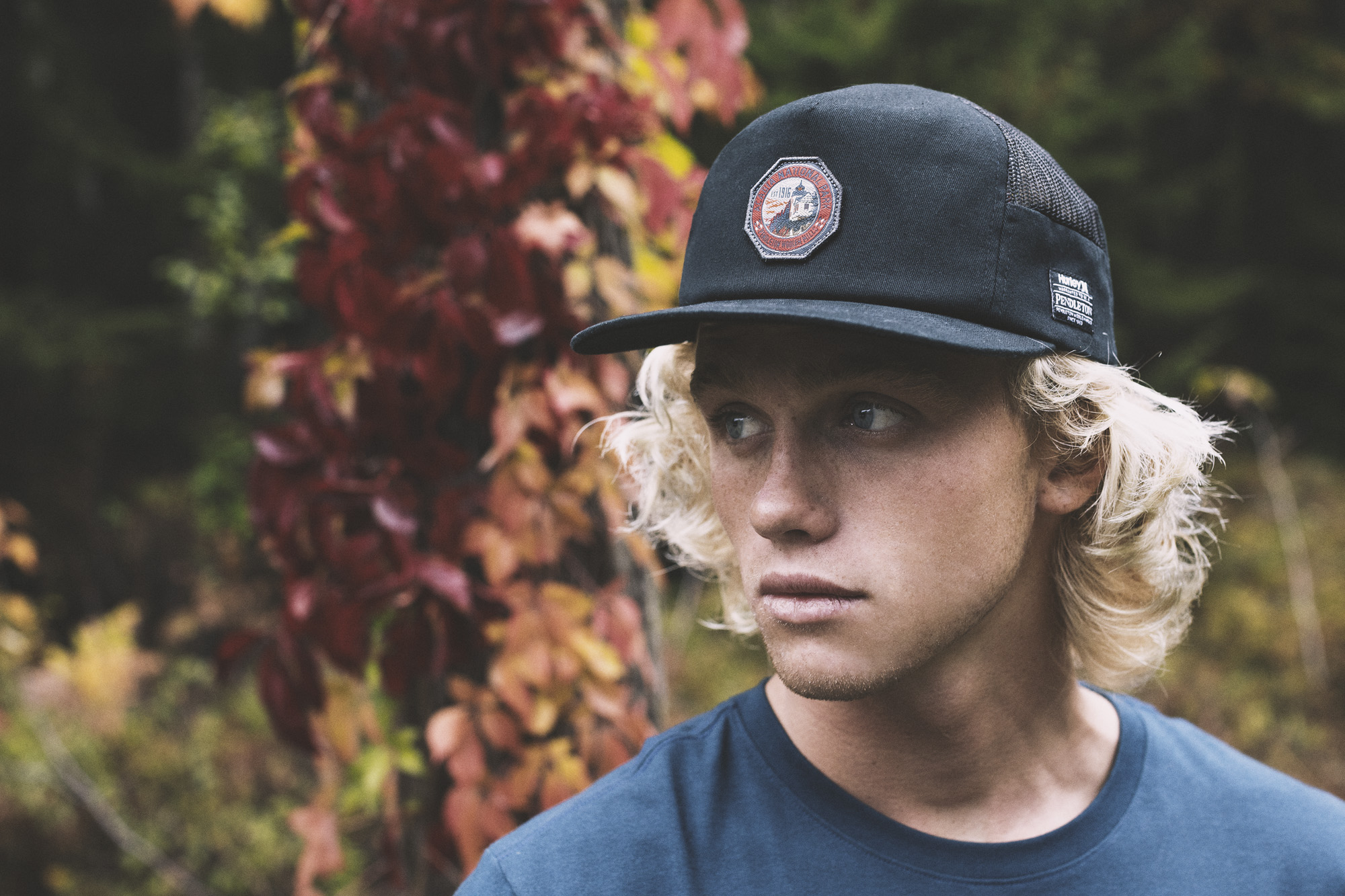 A young man stands in front of an ivy-covered tree, wearing a Pendleton x Hurley baseball cap.