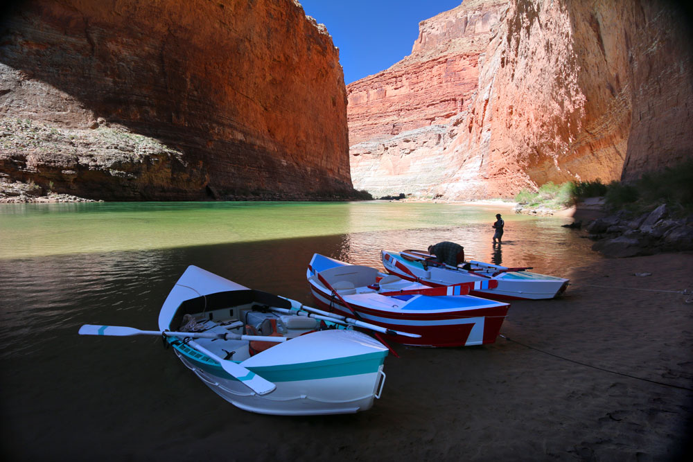 Three beautiful wooden drift boats want in the shallows next to the Colorado RIver, in the shadow of the walls of the Grand Canyon.