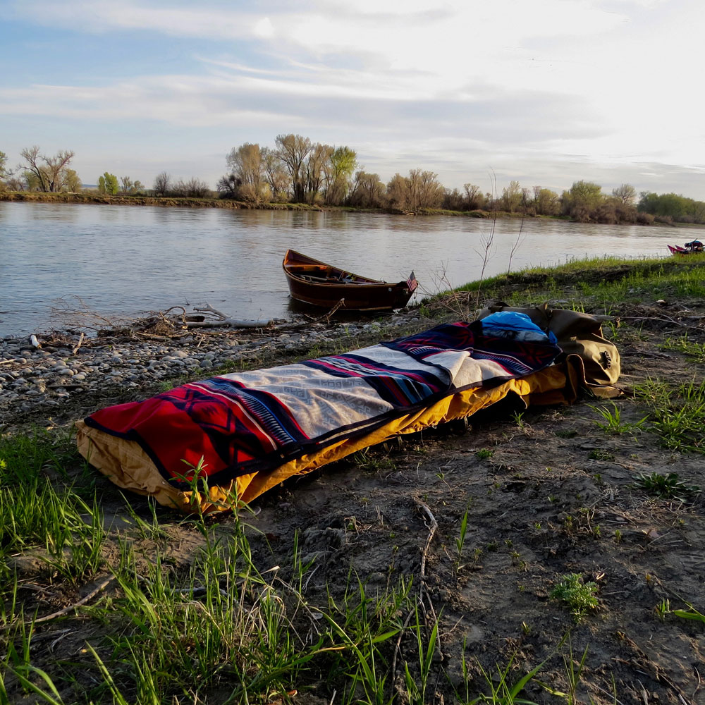 A camp cot set up on the banks of a river, with a wooden boat on the river, and a Pendleton Bighorn blanket on the bed.