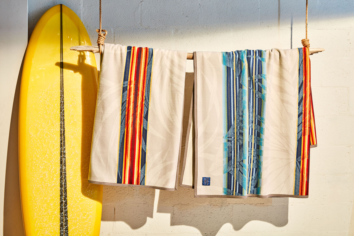 two Pendleton and Tommy Bahama collaborative serape blankets hang from a piece of driftwood next to a yellow surfboard.