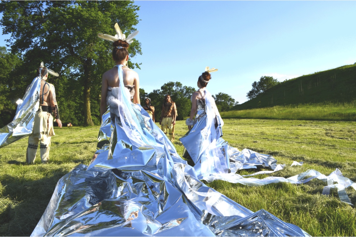 A group of models stand in a meadow, modeling wearable artworks that incorporate long sheets of shining silver Mylar, by artist Wendy Ponca.