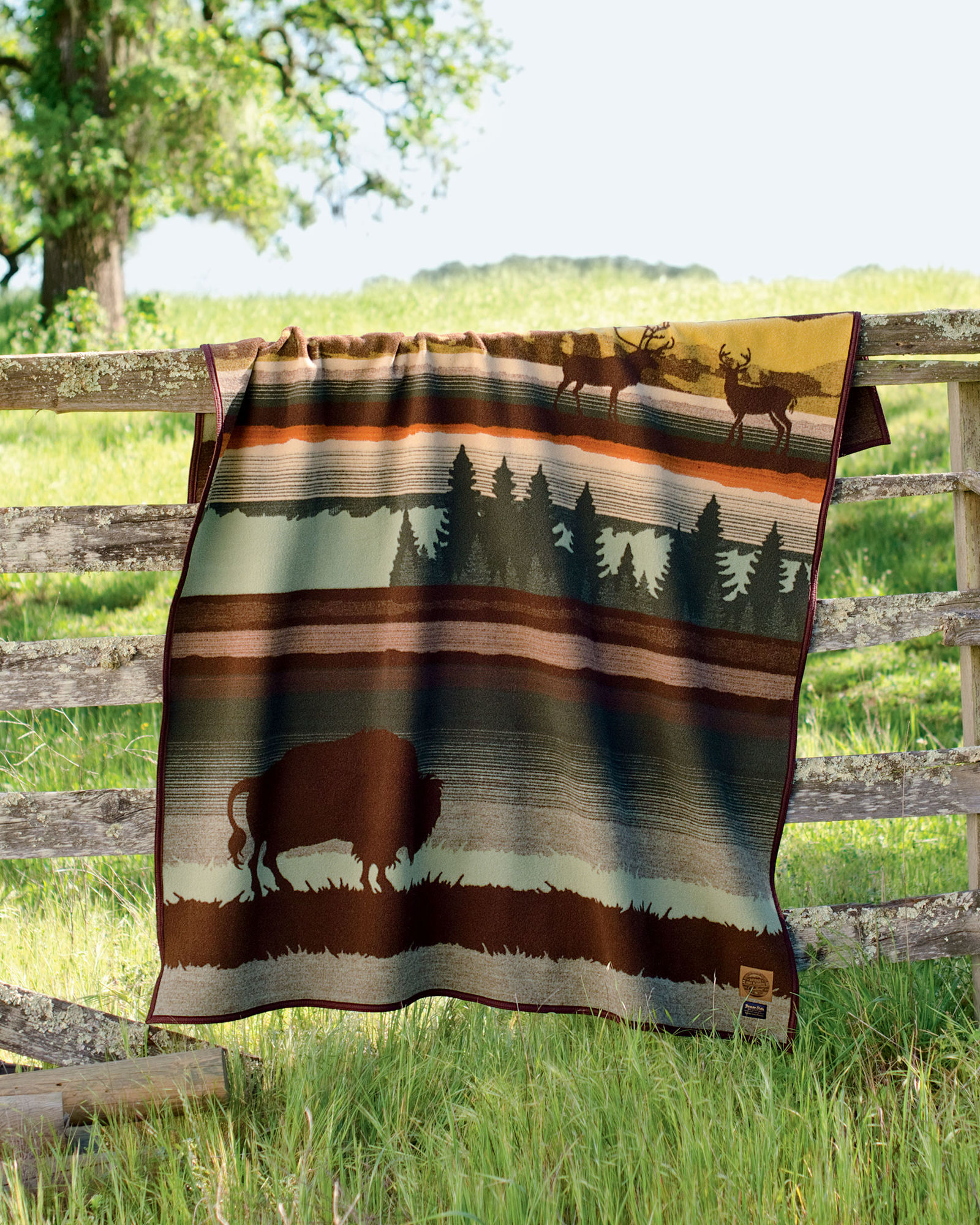 The Buffalo Wilderness blanket by Pendleton hangs over a wooden fence beside a pasture full of bright green grass.