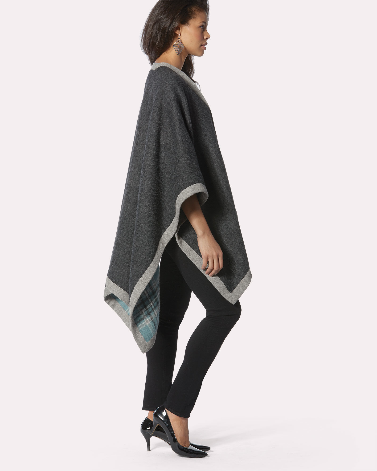 A model stands in profile, wearing a reversible cape of alpaca wool. One side is blue and grey plaid, the other is charcoal grey, with a light grey binding.