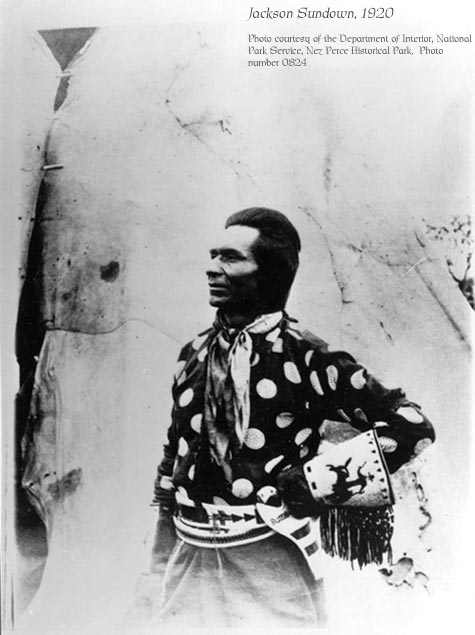 Jackson SUndown poses in front of his family teepee, wearing a dark cotton shirt with large light polkadots, no hat, a beaded belt, and beaded gloves that show a rider on a bucking horse.