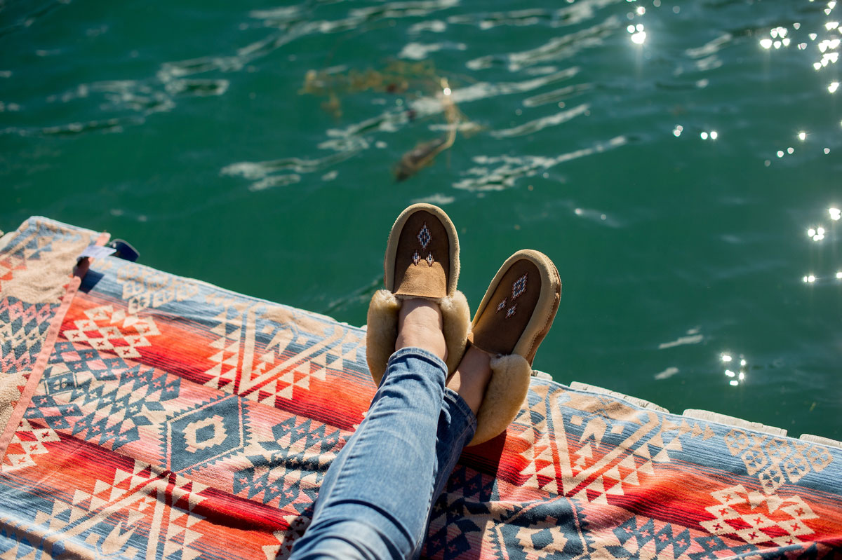 A Pendleton Canyonland beach towel spread on a lake. A woman's lower legs and feet, clad in jeans and beautiful Manitobah moccassins, extend over green lake water that shines with pinpoints of sunlight.