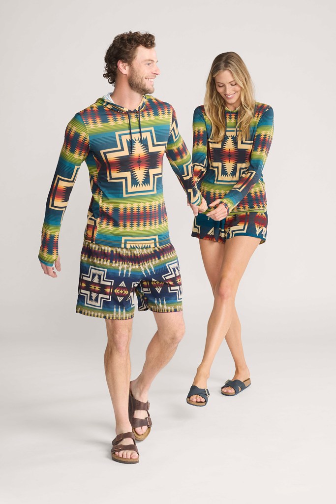 A man and woman hold hands. Both are wearing shorts and rash guards from Pendleton Swim.