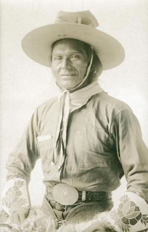 Jackson Sundown smiles for the camera, wearing a chambray shirt, his hat and kerchief, a champion belt buckle, and his beaded gloves.