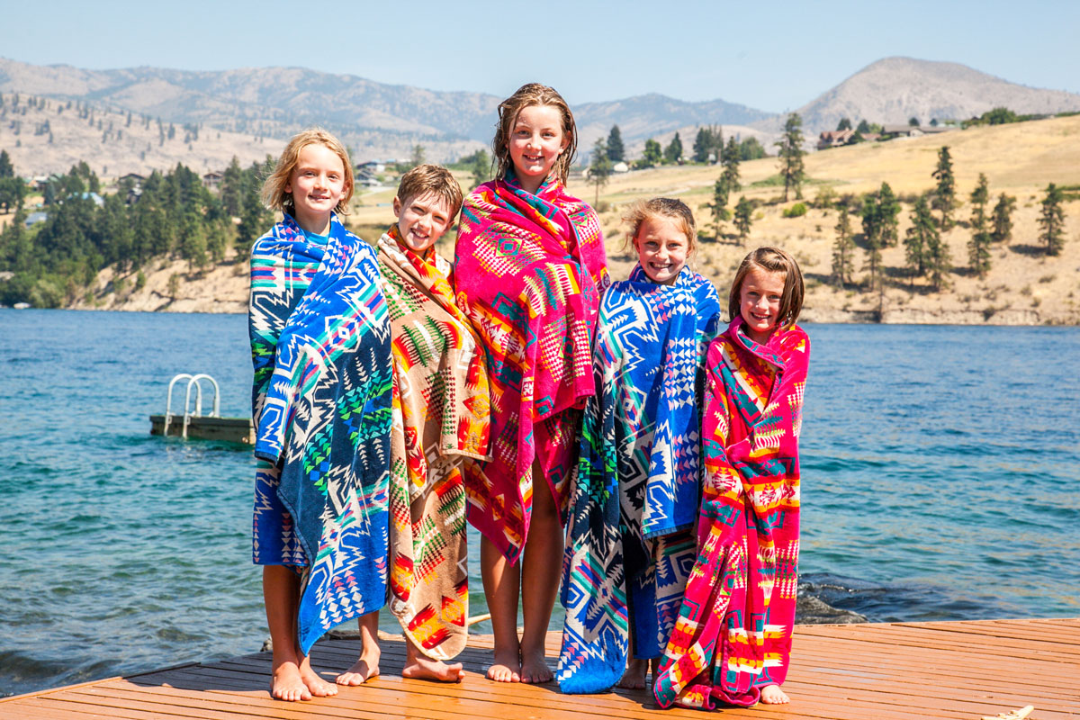 A group of children stand on a dock by a lake, all wrapped in colorful patterned Pendleton beach towels.