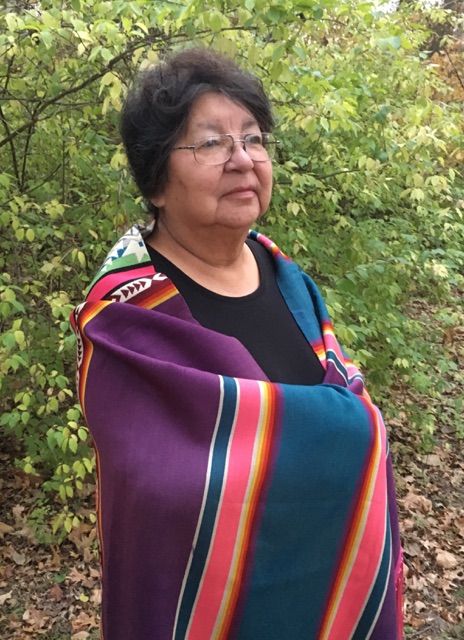 A Native American woman stands in the woods, wrapped in a striped Pendleton blanket.