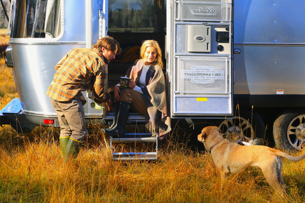 A man and woman at the steps to the Pendleton Airstream, while a dog looks on