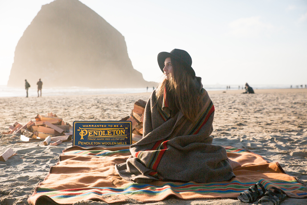 A woman sits on the beach in front of Haystack Rock, wrapped in a Pendleton Camp blanket.