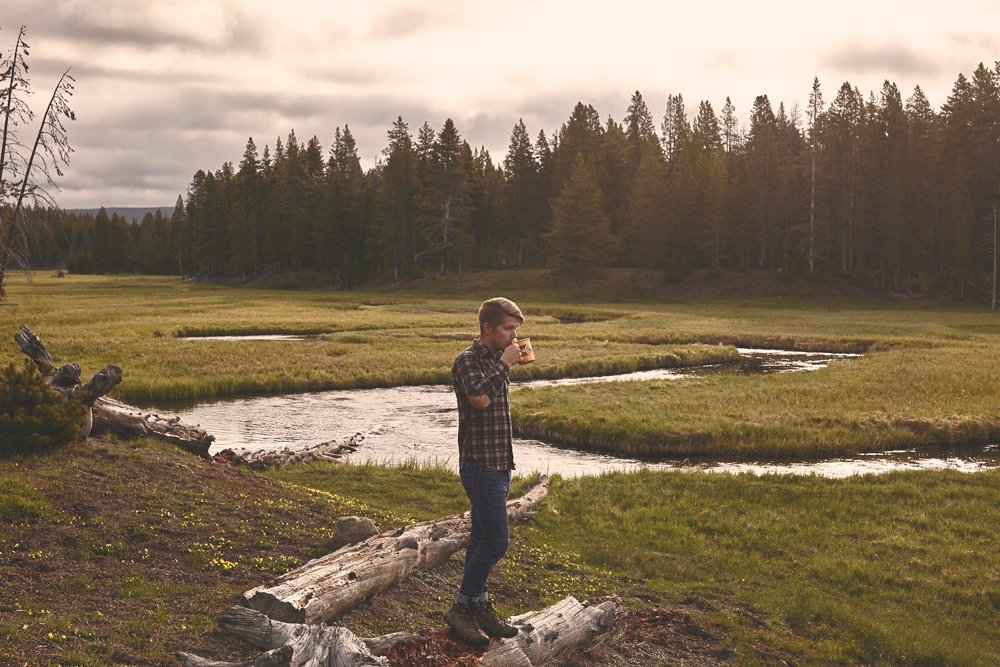 OurFreeWays_ A man wearing a Pendleton shirt drinking coffee by a creek