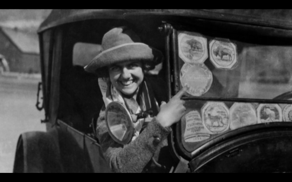 A woman in a Model T points to her windshield stickers.