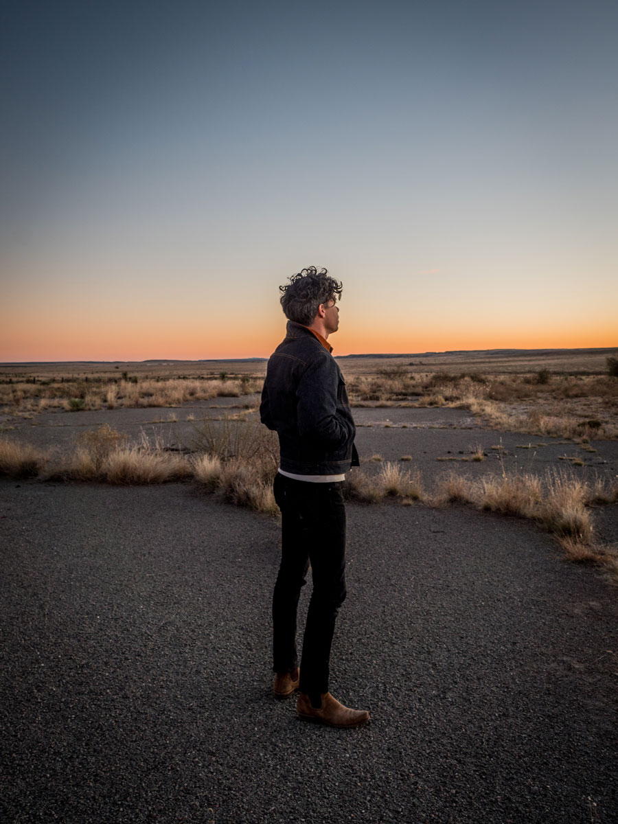 A man stands in the desert, the horizon glowing with either sunrise or sunset, wearing Ginew denim jeans and jacket.