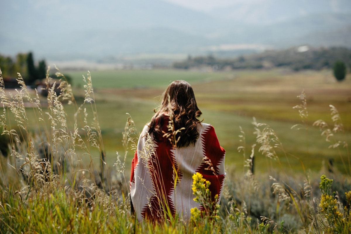 A woman sits, wrapped in a Pendleton Brave Star blanket, in a mountain meadow. Her back is to the camera. 