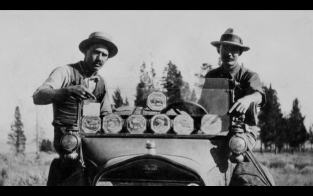 1920s men point to their car's windshield which is adorned with large National Park stickers. 