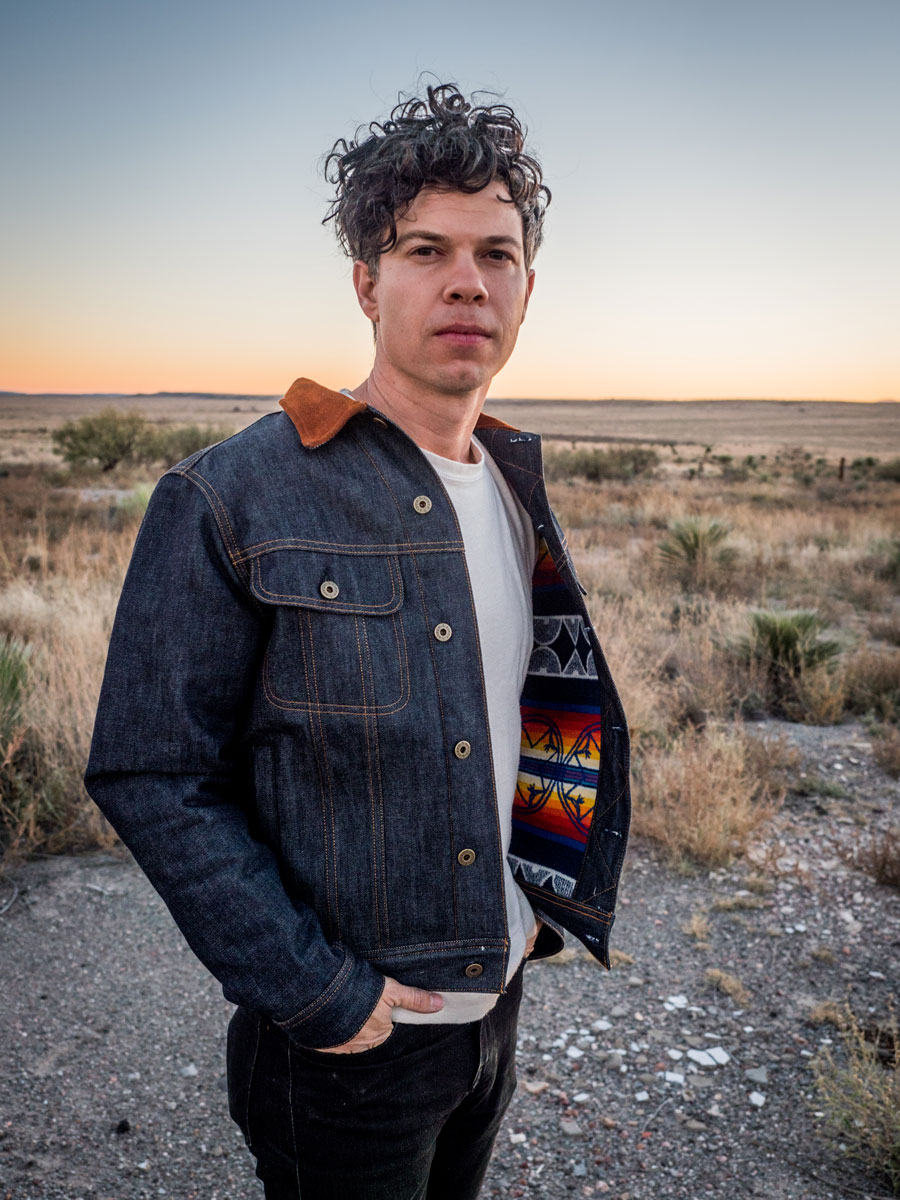 A man stands in a deserted field wearing a Ginew x Pendleton denim jacket.