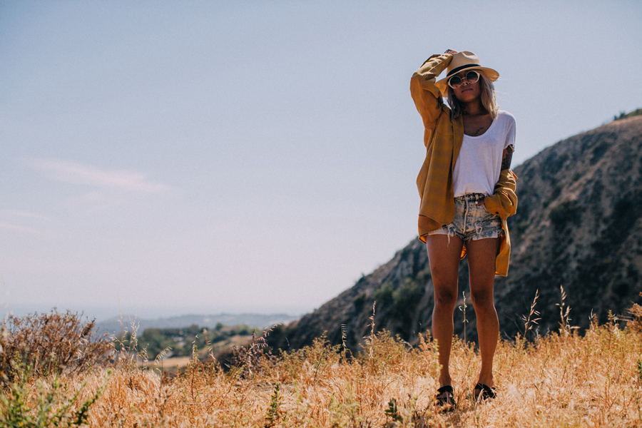 A young woman stands on a hillside, wearing a gold Pendleton shirt and a Pendleton hat.