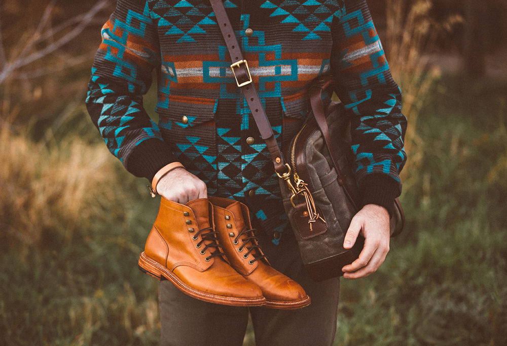 A man stands in the woods holding a pair of boots and holding a bag, wearing a Pendleton blanket coat. 