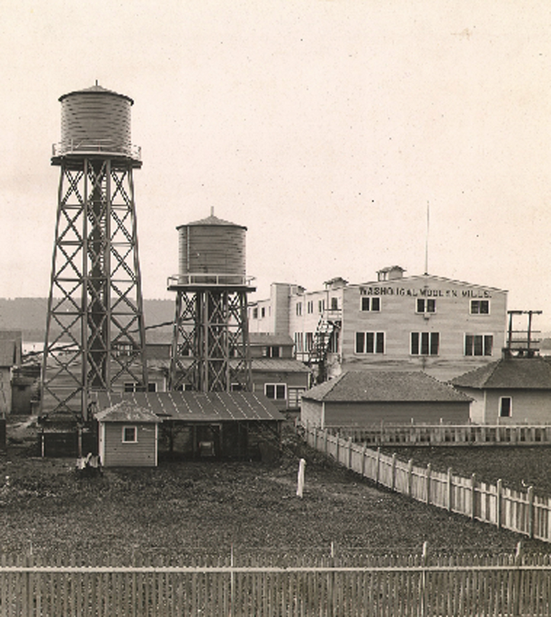 A vintage sepia-toned photo of the Washougal woolen mill owned by Pendleton Woolen Mills. The mill is two stories tall and in the photo, it is dwarfed by two water towers.