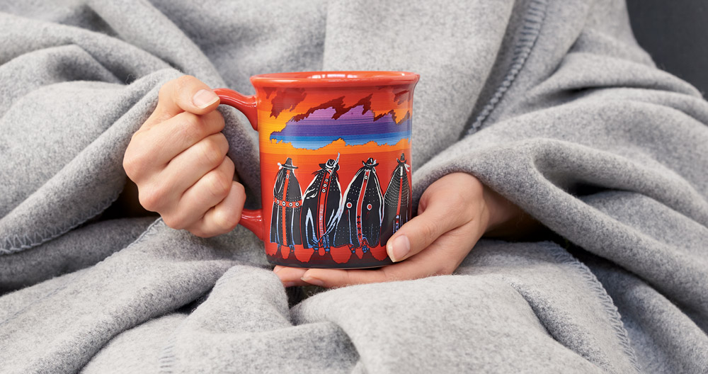 A blanket-wrapped woman's hands holding the Rodeo Sisters mug.