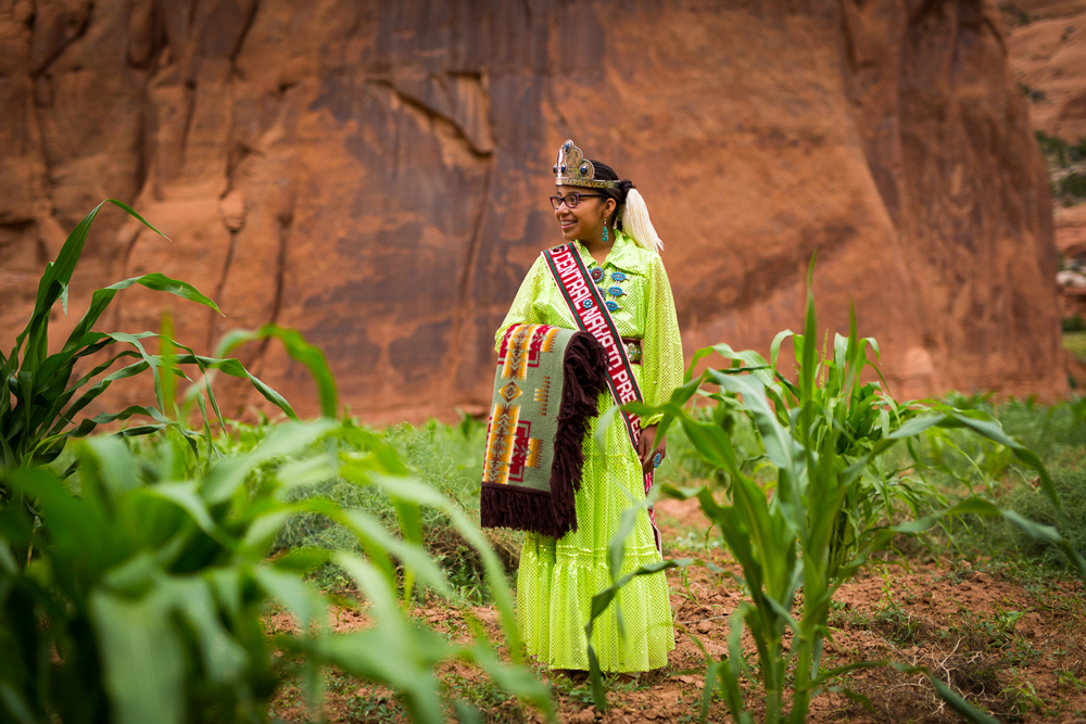 Tonisha Draper stands in her family's corn plot, ed rock behind her. She is wearing a bright green traditional Navajo gown, a crown, and she is holding a fringed Pendleton robe in the Chief Joseph pattern. 