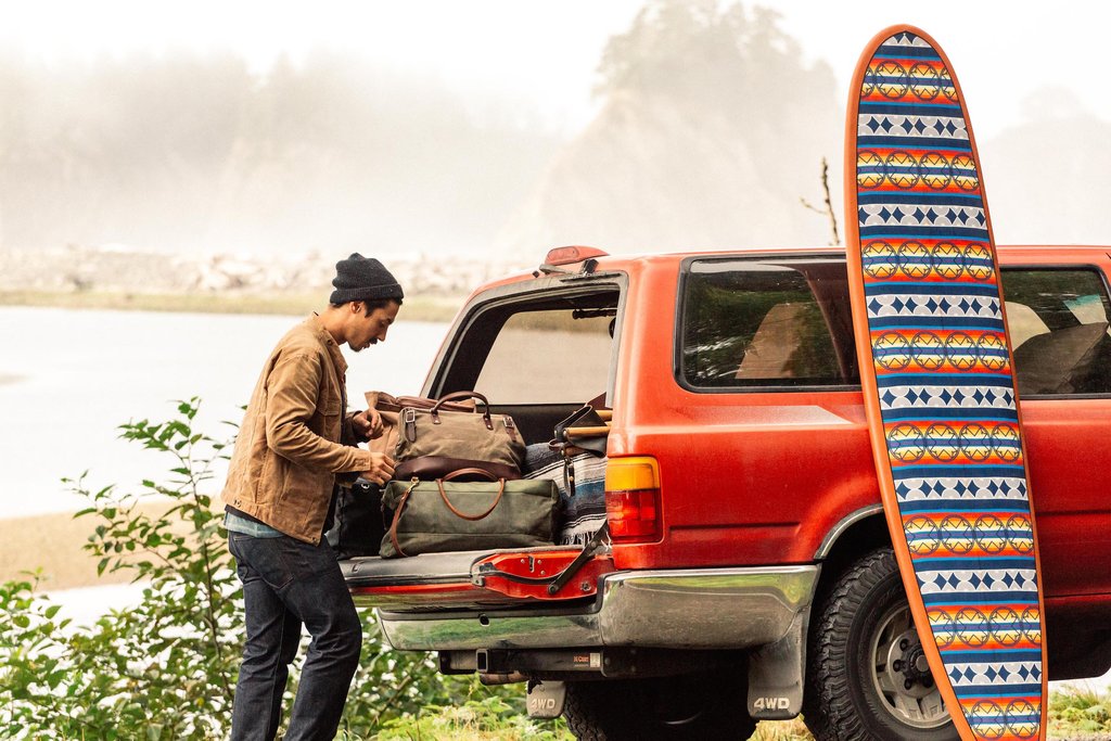 A man unpacks the back of his rig, and a Ginew surfboard with a Pendleton pattern leans against the vehicle.