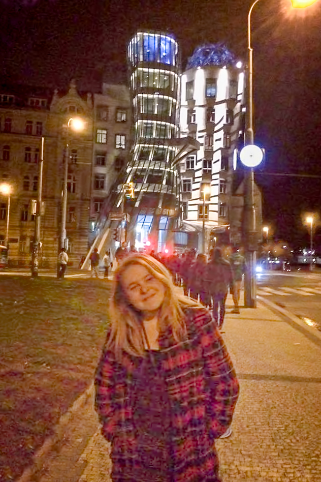 A young woman in a vintage Pendleton coat stands before Prague's Dance House.