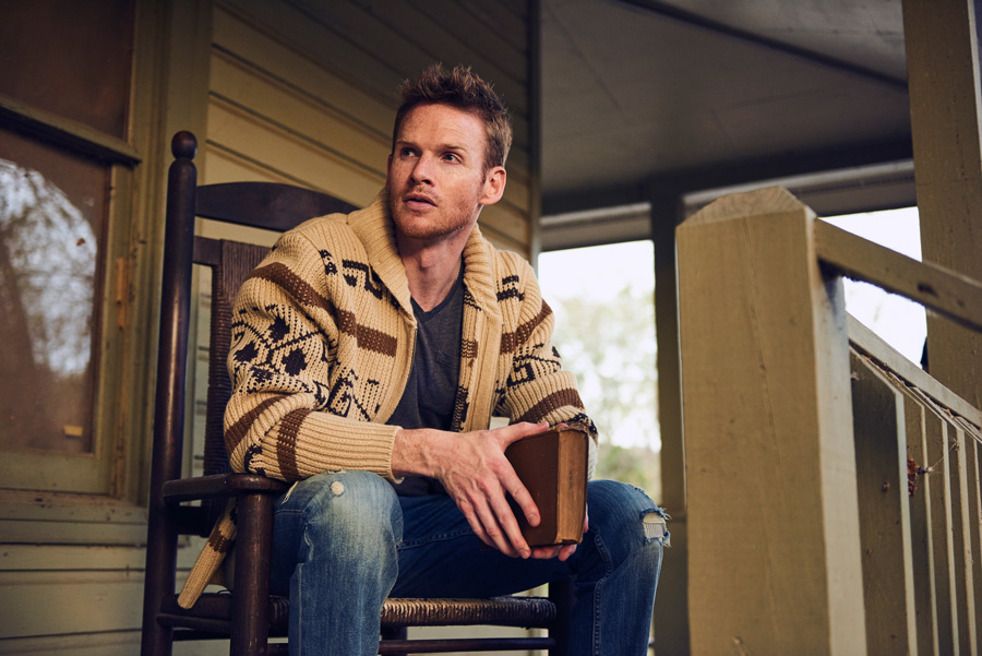 A man sits in a rocking chair on the porch of an oder home, holding a book and wearing a Pendleton Westerley cardigan sweater.