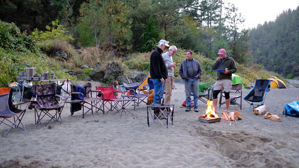 A group of river rafters eat breakfast on a riverbank, near a fire.