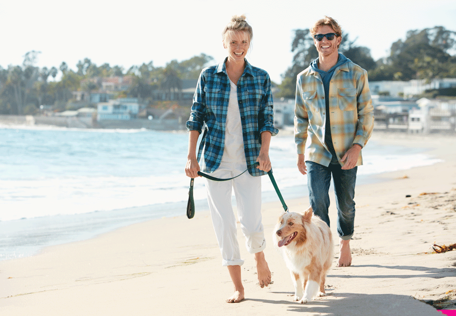 A man and woman walk down the beach with a dog, wearing Pendleton Board Shirts.