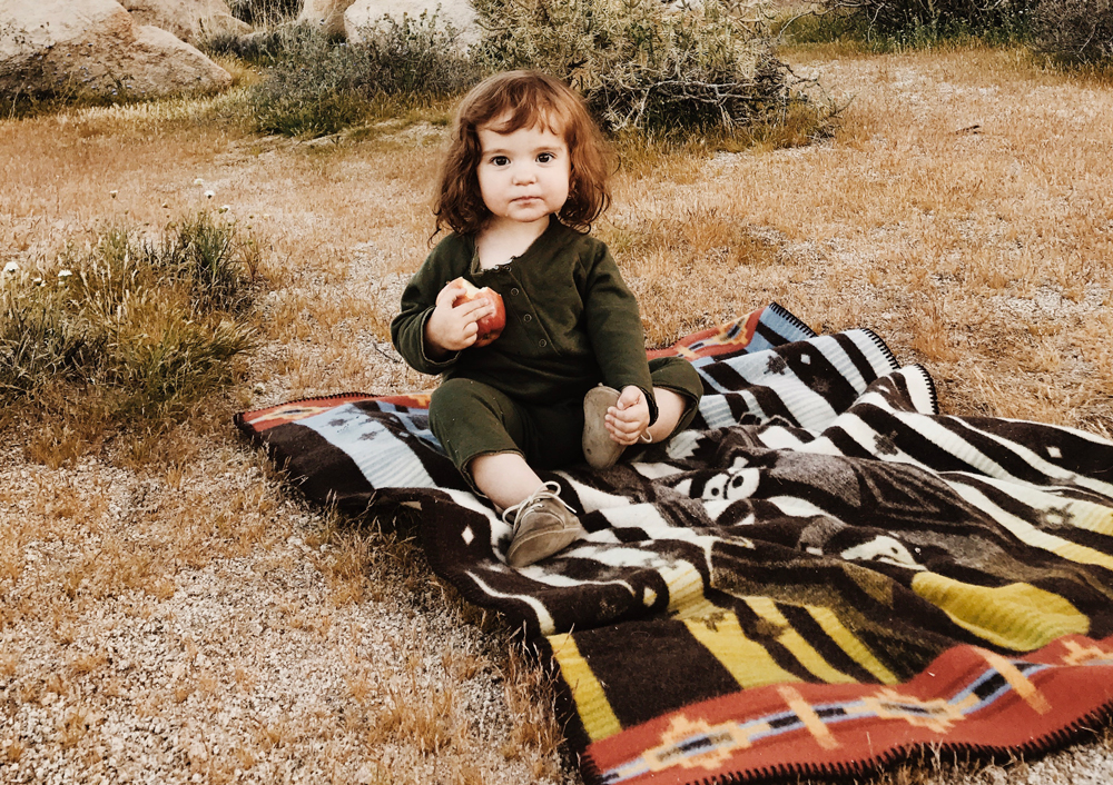 A little girl hoding an apple sits on a Pendleton blanket. 