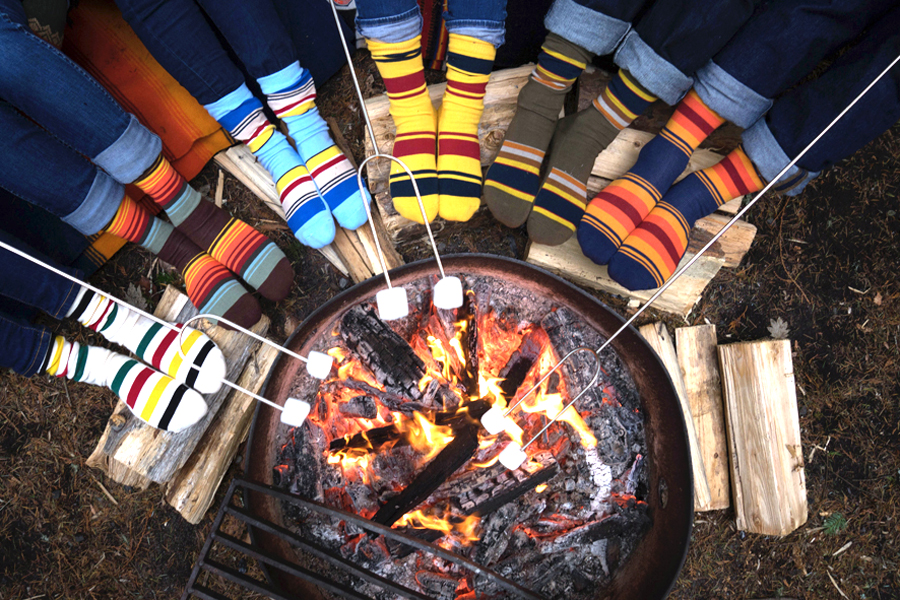 A campfire, over which marshmallows are being toasted, and six people's feet wearing Pendleton socks. 