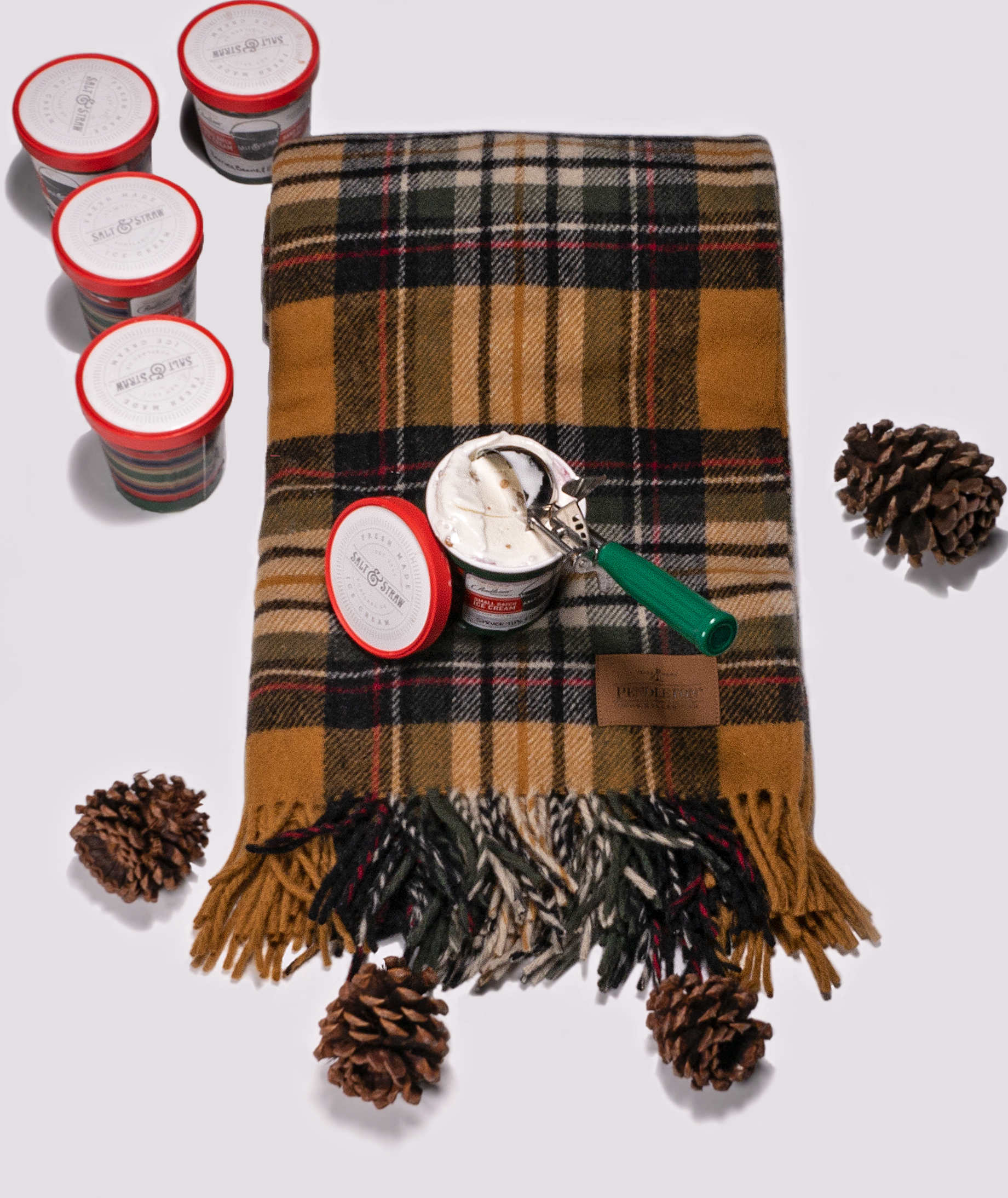 A folded Pendleton Motor Robe in a plaid of gold, black, tan, white and red sits on a white background, next to four pinecones. Four slosed pint containers of Salt & Strwa ice cream are to the upper left of the blankets, and one open pint of ice cream sits on the throw, with a green-handled ice cream scooper in the ice cream. 