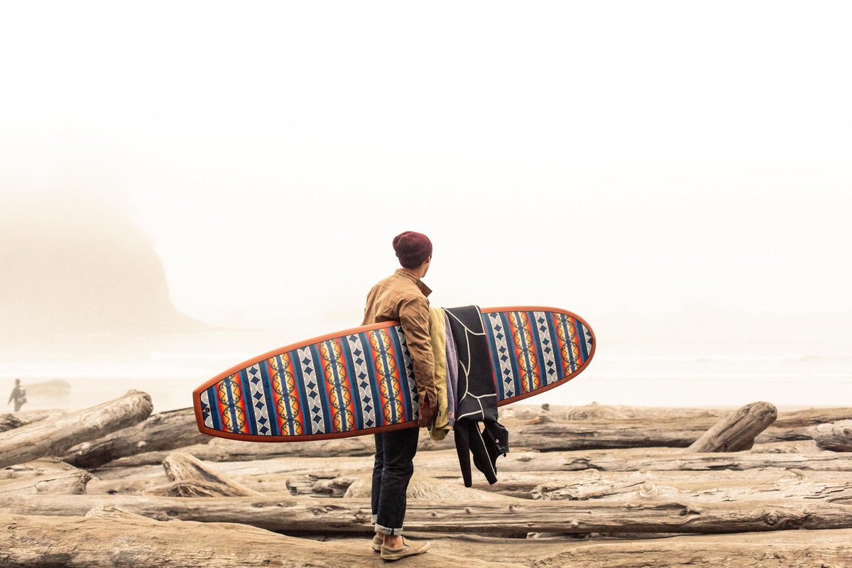 A man stands on the beach, holding a Ginew surfboard.