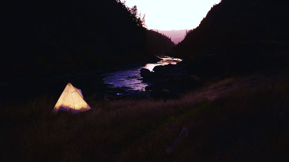 A lit canvas tent glows beside a river in Oregon, as the sun sets.