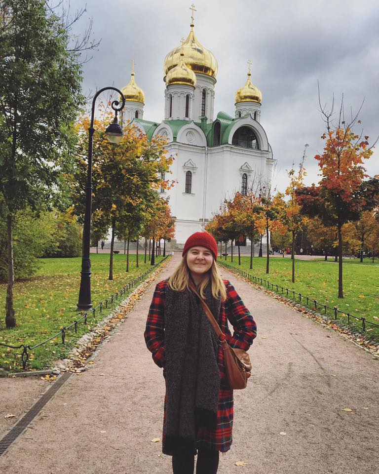 A young woman in a Pendleton coat stands before a church with gold onion domes.