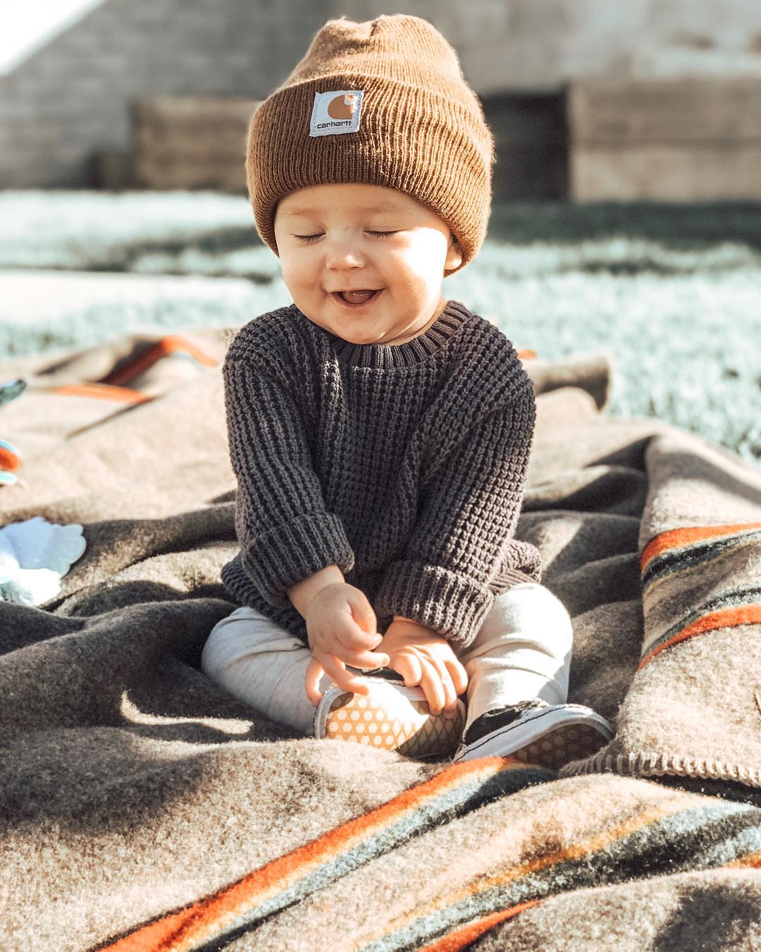 A smiling baby wearing a knit sweater, cap, track pants and small baby tennis shoes sits on a Pendleton camp blanket in the sunshine.