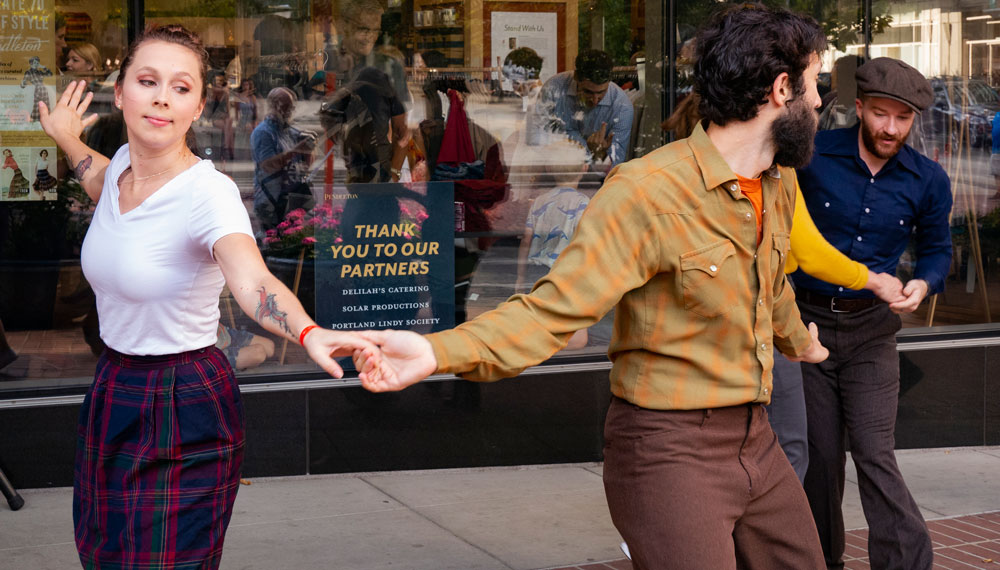 Linday Hop dancers dancing on sidewalk in front of pendleton store, one woman, two men