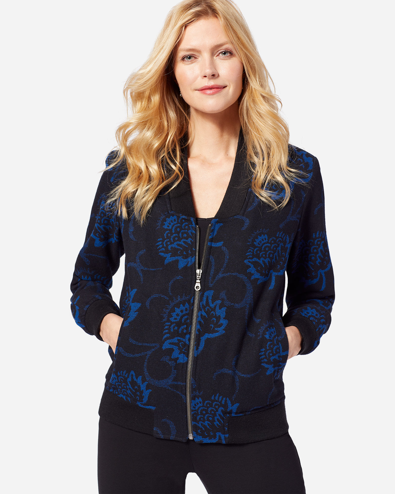 A woman with long blond hair stands in front of a white background, wearing black pants and a black wool zip-up jacket with blue thistle flowers in the fabric pattern.