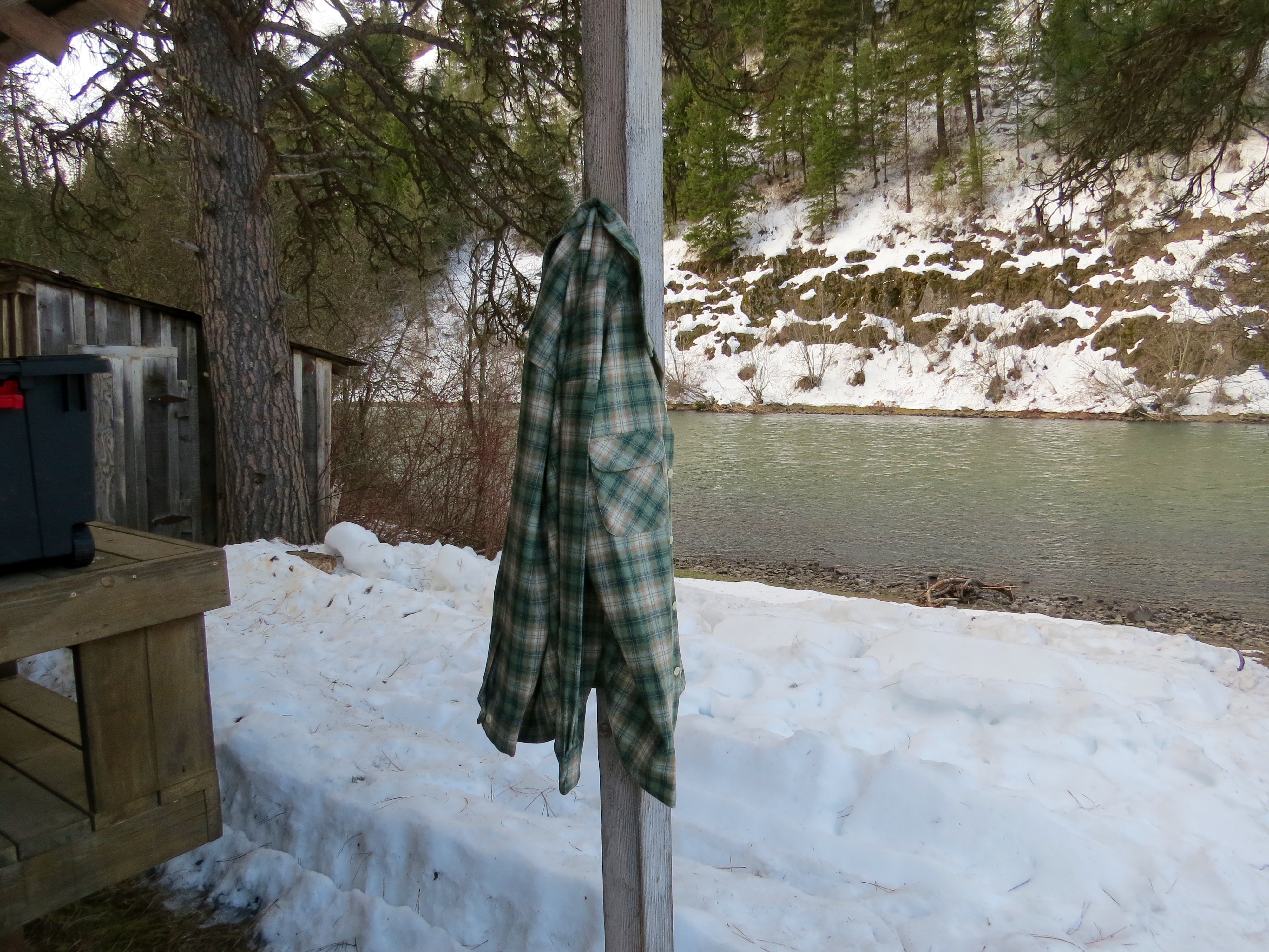 A plaid wool Pendleton shirt hangs on a pole near a snowy riverbank. 