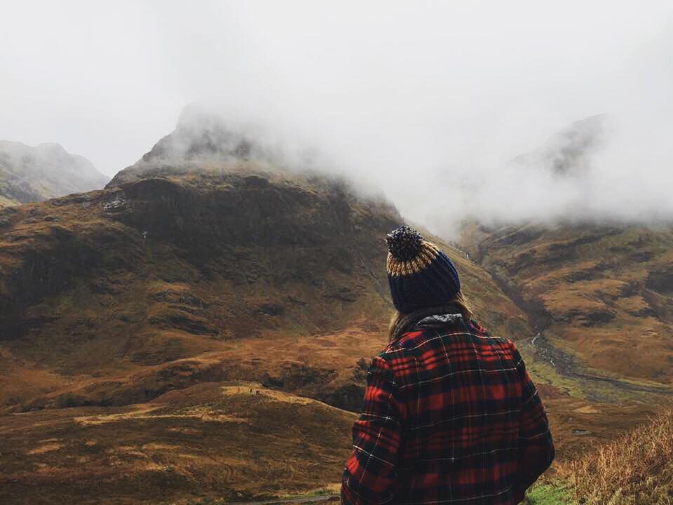 A young woman in a Pendleton coat stands looking at the Three Sisters peaks in Glencoe, Scotland.