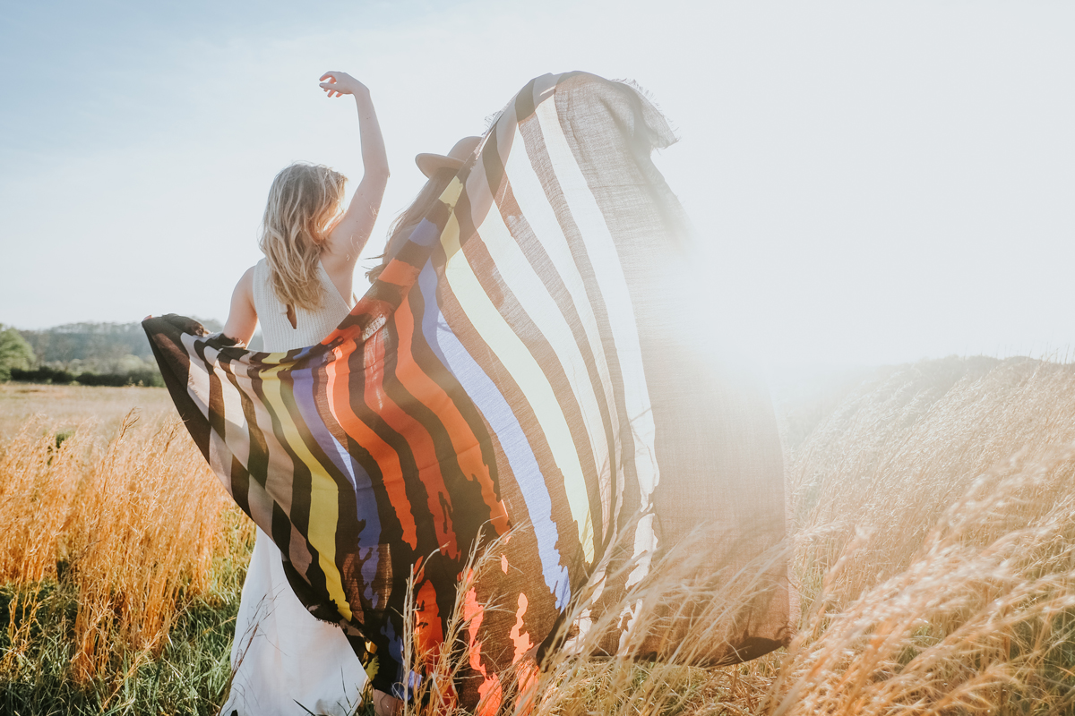 Two women in a meadow raise their arms and a Pendleton scarf to the sun. 