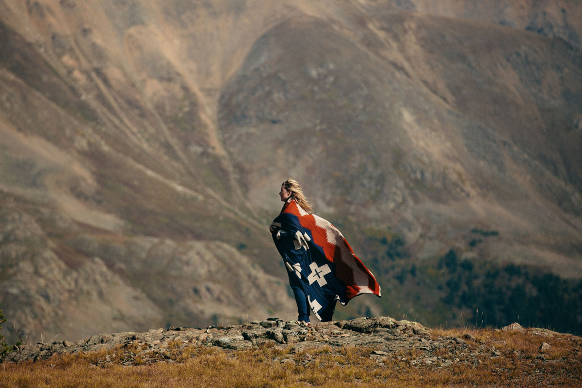 A young woman stands in front of a mountain, wrapped in a Pendleton Brave Star blanket.