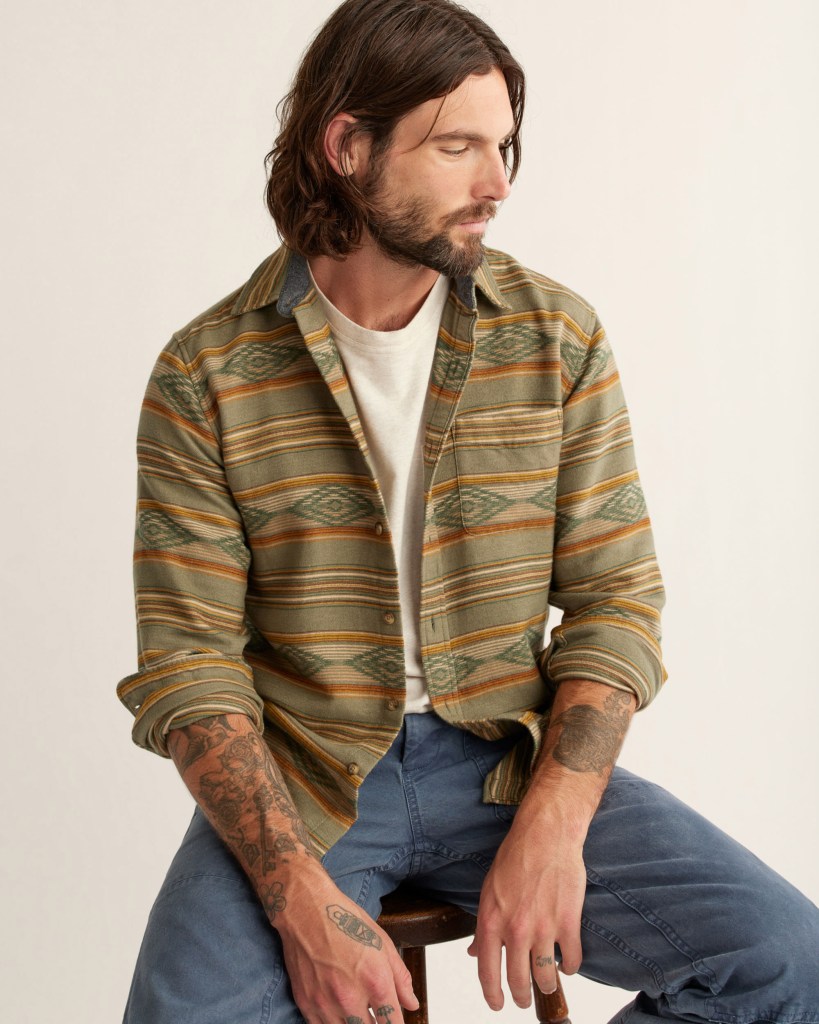 A young man with dark hair and a beard sits on a wooden stool, wearing the Marshall Overshirt by Pendleton.