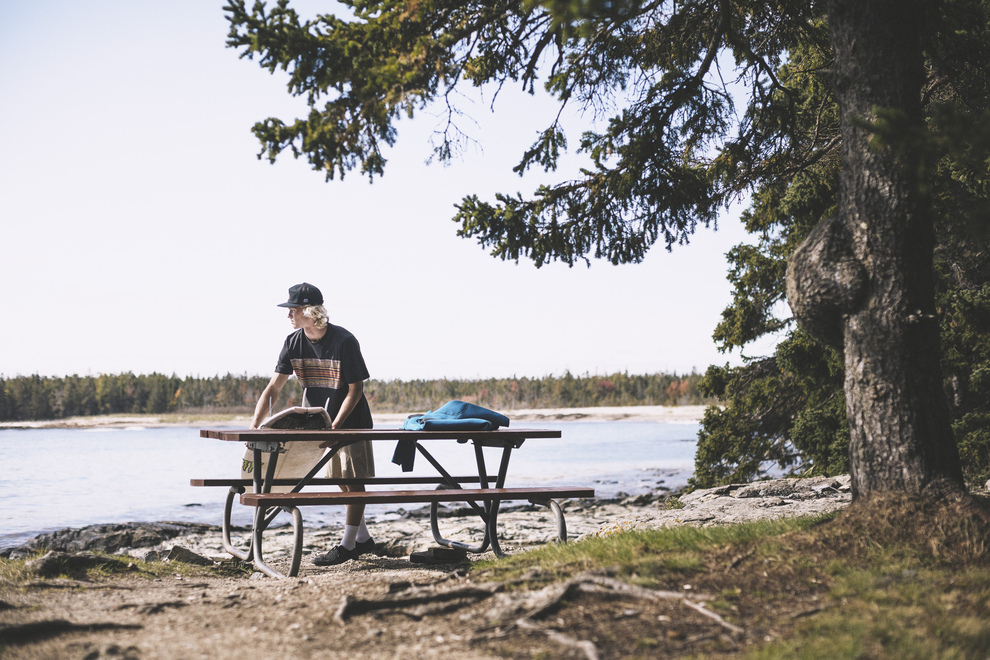 A young man leans on a wooden picnic table by the shore of a lake, wearing Pendleton x Hurley activewear.