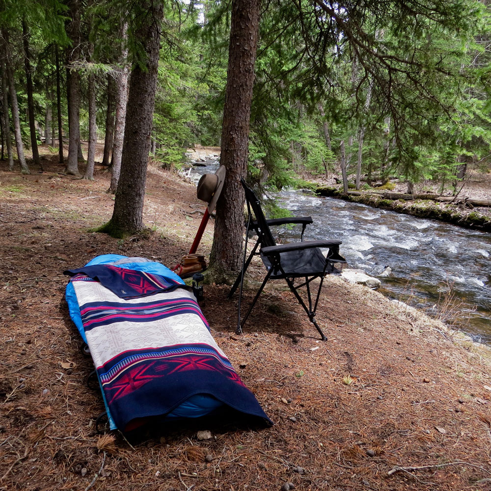 A camp cot and folding camp chair on the banks of a rushing river, under evergreen trees. I Pendleton Bighorn blanket is on the cot.