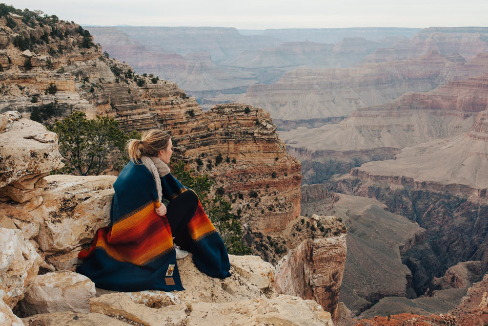 A young woman wrapped in a Pendleton Grand Canyon National Park blanket sits on the Grand Canyon's rim, looking into the distance.