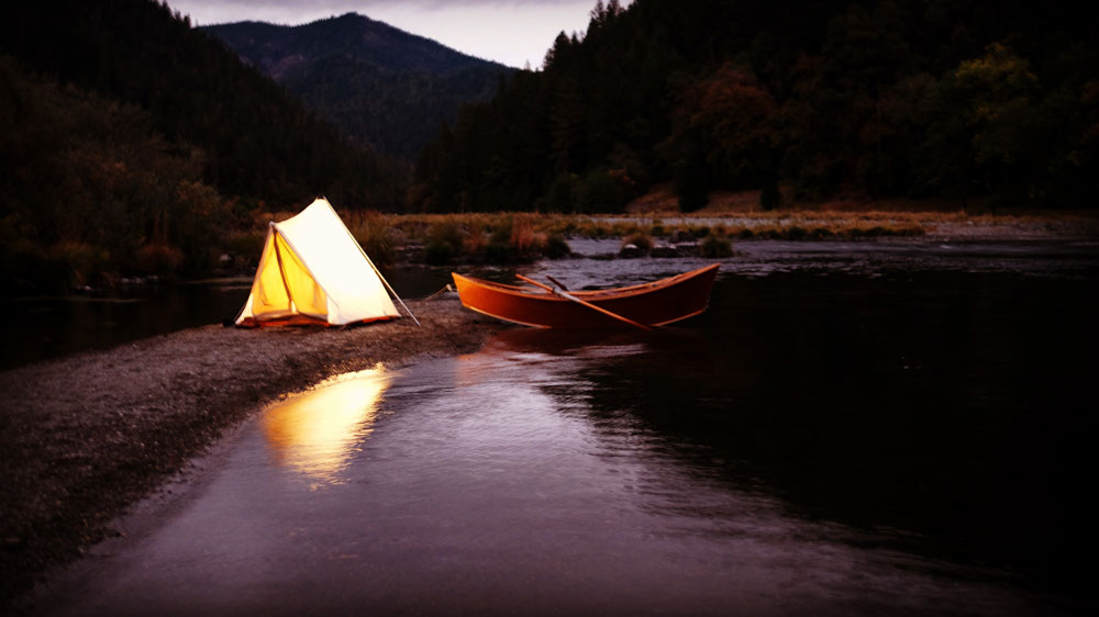 An evening outdoors shot, with a lit canvas tent, and an empty wooden drift boat by the shore of a small river.